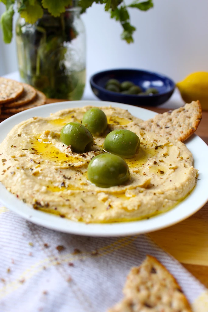 Addicting and Easy Mediterranean Hummus spread onto a plate. Delicately topped with beautiful bright green castelvetrano olives, and drizzled with olive oil.