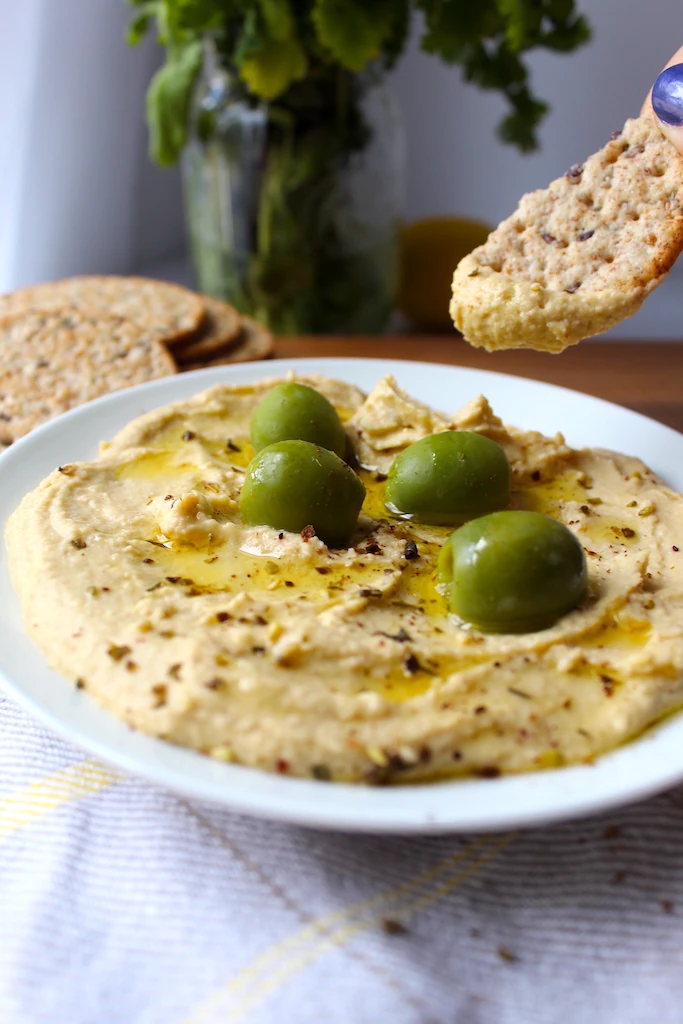 Creamy hummus is being scooped up by multigrain cracker broken in half. 