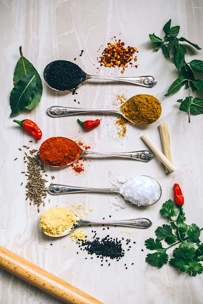 Five spoons full of beautiful spices and flavors are pictured for use of Portuguese Cuisine. Peppers, herbs and spices are bountiful and spilling over onto the counter.