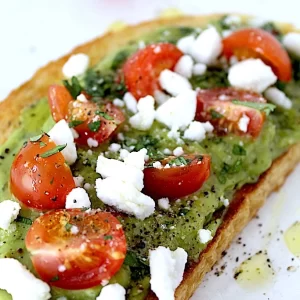 Spicy Avocado Toast with bright red, juicy, sliced tomatoes, white cotija cheese crumbles and spicy avocado spread onto a thick toasted slice of bread.