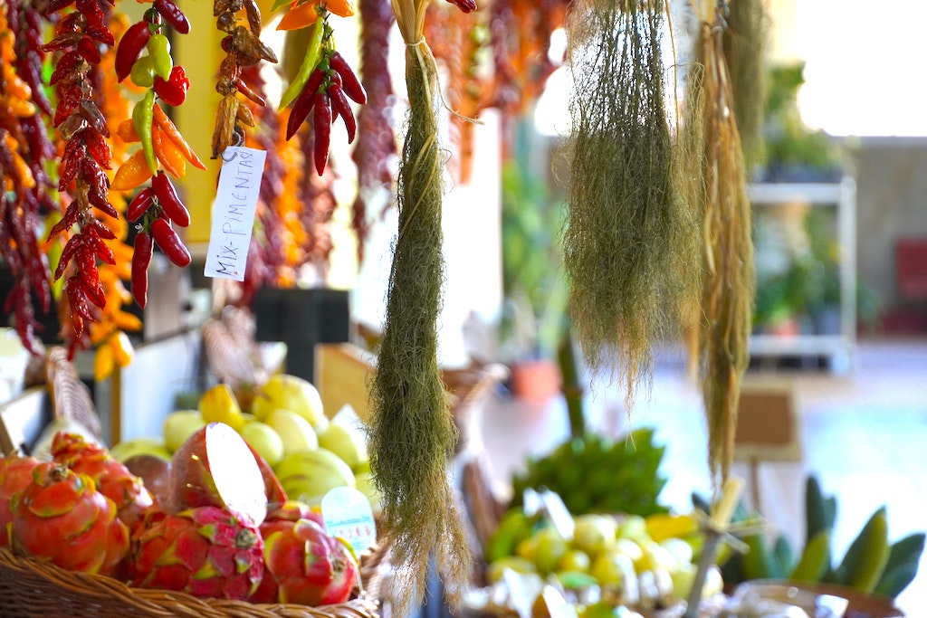 Dried herbs hang beside a string of chilis. Various fruits and veggies are overflowing from baskets on display. Food is sold for Portuguese Cuisine at an open air farmers market. The Mercado dos Lavradores in Funchal, Portugal.