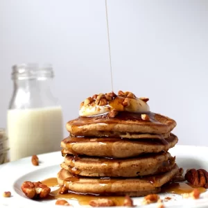 Pouring golden maple syrup over the top of gorgeous, light and fluffy pumpkin spiced pancakes surrounded with pecans and topped with a dollop of yogurt.