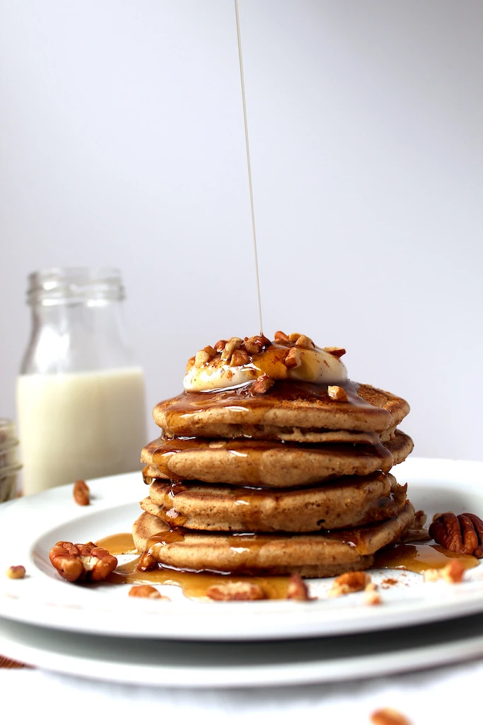 Pouring golden maple syrup over the top of gorgeous, light and fluffy pumpkin spiced pancakes surrounded with pecans and topped with a dollop of yogurt.