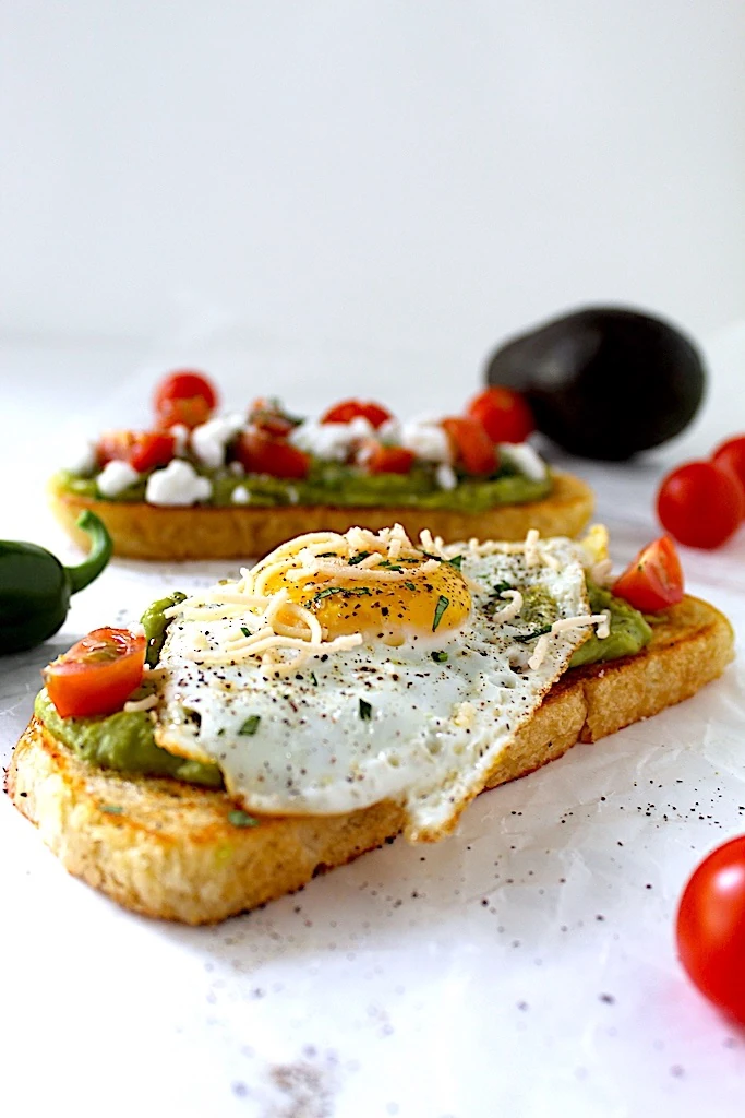 Spicy yet creamy avocado toast covered with tomatoes, parmesan, parsley, and olive oil. Garnished with an elegantly cooked sunny-side-up egg!