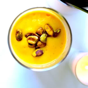 A picture of a Cardamom Spiced Mango Lassi. Thick, creamy, frothy, yogurt-based drink blended with mango, cardamom, and sweetener. Delicately topped with pistachios.