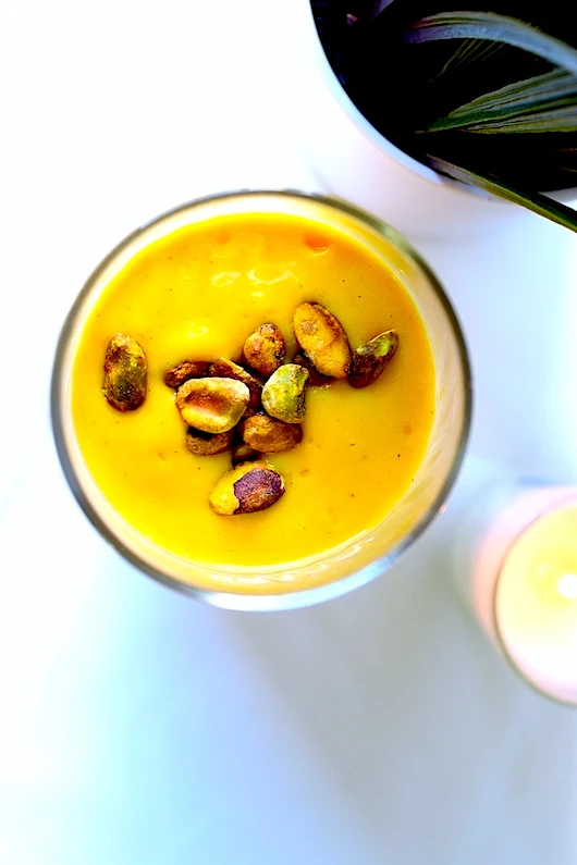 A picture of a Cardamom Spiced Mango Lassi. Thick, creamy, frothy, yogurt-based drink blended with mango, cardamom, and sweetener. Delicately topped with pistachios.