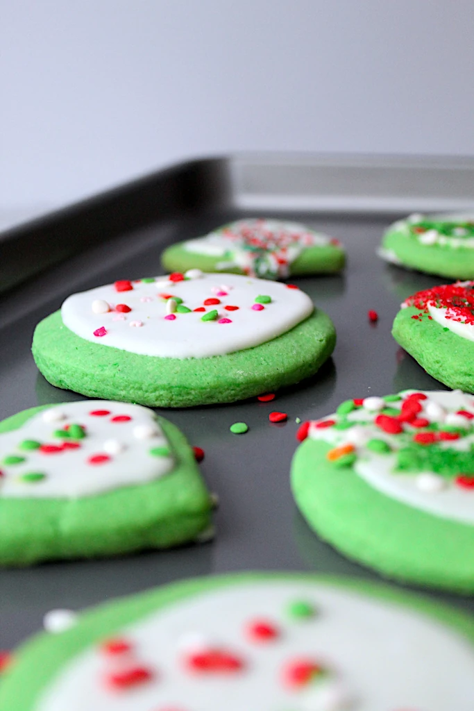 A cookie sheet lined with sprinkles covered, bright green Grinch Inspired Almond Sugar Cookies and frosty white royal icing.