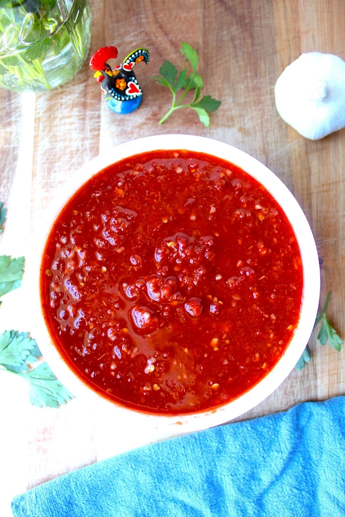 Garlic, fresh herbs and a Portuguese rooster surround a bowl of brilliantly red Vinha D'alhos Portuguese Marinade.