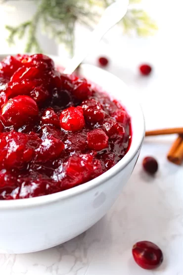 A strikingly white bowl holds brilliant, vibrant, red Easy Sugared Cranberry Sauce. Christmas Holly and cinnamon sticks fade into the background.