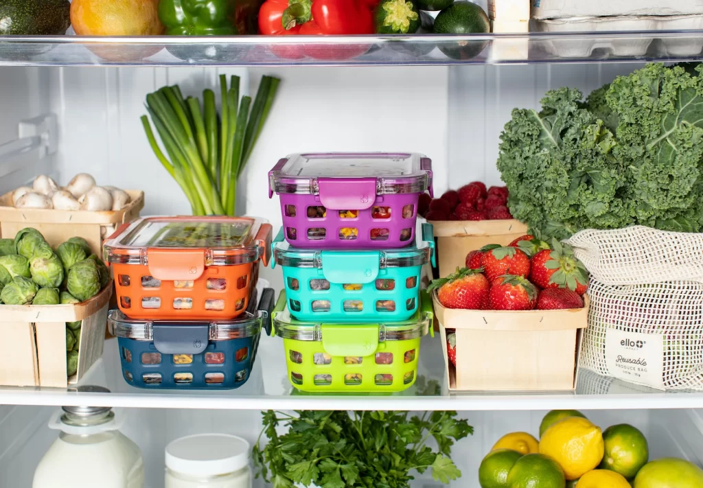 An open fridge reveals a stack of colorful Tupperware, fresh veggies and various fruits as well.