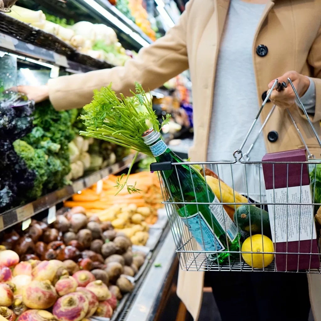 A woman in a tan coat is shopping for groceries using a basket. She is reaching for some fresh vegetables and Save Money on Groceries.