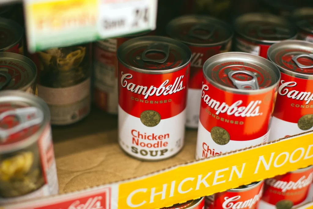 A close-up of a box filled with soup cans colored in red and white. Prices are blurred but are shown for people Save Money on Groceries.