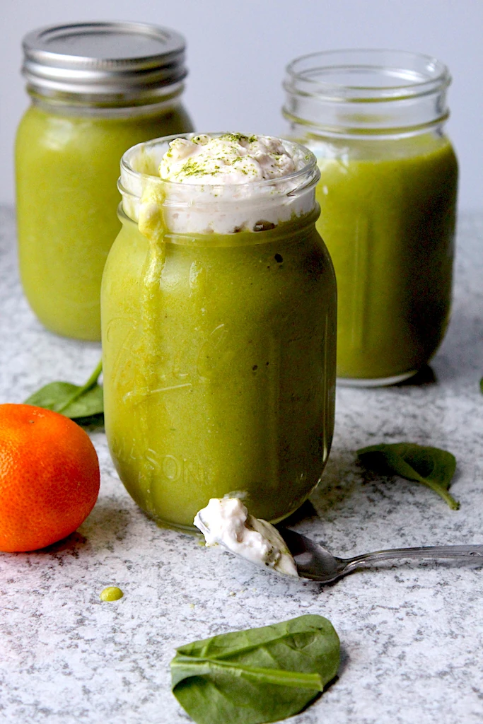 Three mason jars are filled with beautiful Green Matcha Citrus Smoothie mixture, dripping down the side of the jars. The smoothies are topped with almond whipped cream and sprinkled with gorgeous green matcha powder.