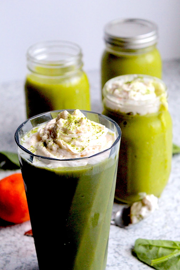 Displaying a beautiful Green Matcha Citrus Smoothie. Multiple glasses shown with dramatic shadow overcast, an orange and some scattered spinach are faded into the background on the granite counter top. The smoothies are topped with fluffy, white whipped cream and gorgeous matcha sprinkled on top.