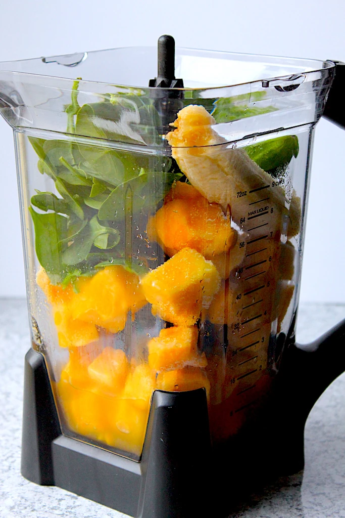 A tall blender full of beautiful and fresh ingredients, green, orange and yellow.