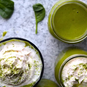 Gorgeous green smoothies, topped with almond whipped cream and sprinkled in matcha powder. Spinach is scattered on the granite counter top surrounding the glasses. A spoon full of almond whipped cream lays beside a full glass of smoothie.