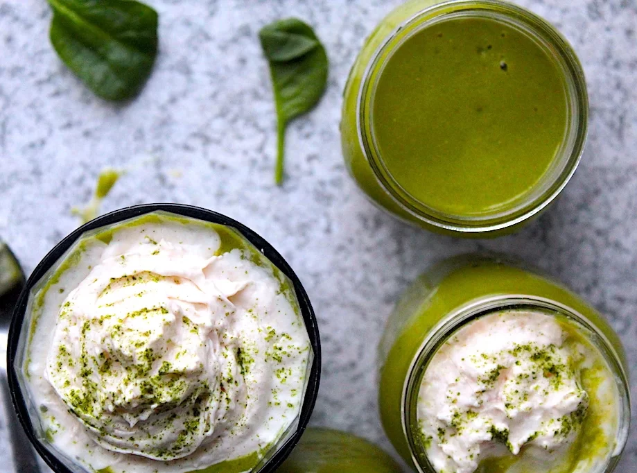 Gorgeous green smoothies, topped with almond whipped cream and sprinkled in matcha powder. Spinach is scattered on the granite counter top surrounding the glasses. A spoon full of almond whipped cream lays beside a full glass of smoothie.