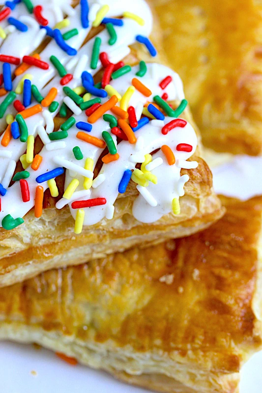 a single puff pastry square, golden and flaky, holds secrets within. A slice reveals a river of fruity jam, while creamy cream cheese crowns the pastry. Colorful sprinkles dance on top, inviting pure delight in every bite.