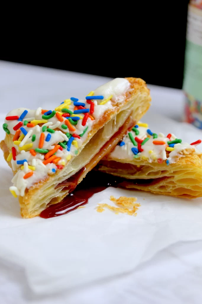 Imagine a stack of puff pastry squares, freshly baked to a golden crisp. With a gentle slice, the layers reveal a tantalizing secret - a rich filling of fruity jam, its deliciousness oozing out invitingly. Crowned atop is velvety cream cheese, a perfect creamy companion to the flaky pastry. A shower of colorful sprinkles cascades down, adding a playful touch to this irresistible masterpiece.