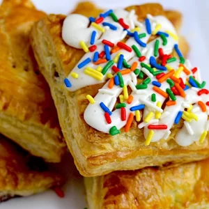 Indulge in a delightful treat - a stack of puff pastry squares, each layer baked to golden perfection. Between the flaky layers rests a burst of fruity jam, adding sweetness and vibrancy. Crowned with velvety cream cheese, offering a creamy contrast, and adorned with a cascade of colorful sprinkles, creating a joyful feast for both the eyes and the taste buds.