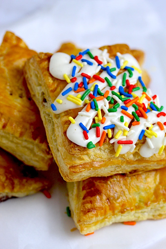 Indulge in a delightful treat - a stack of puff pastry squares, each layer baked to golden perfection. Between the flaky layers rests a burst of fruity jam, adding sweetness and vibrancy. Crowned with velvety cream cheese, offering a creamy contrast, and adorned with a cascade of colorful sprinkles, creating a joyful feast for both the eyes and the taste buds.