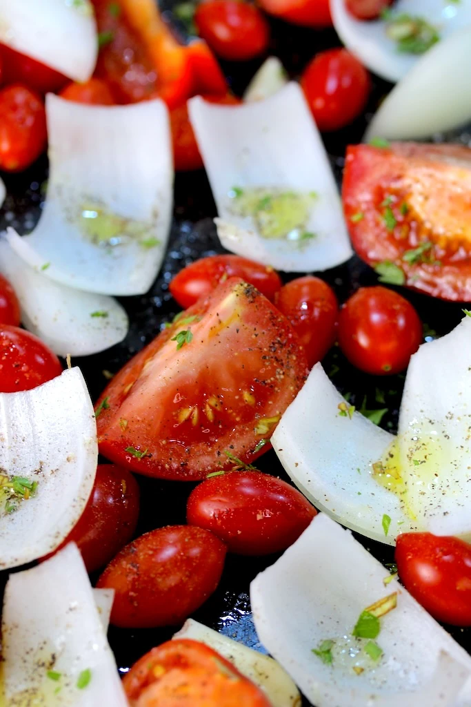 Fresh bright red tomatoes and white onions are peppered with fresh herbs and spices, ready to roast.